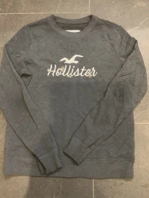 Grå sweatshirt från Hollister - Mysig grå sweatshirt från Hollister med broderad logga och text på bröstet i nytt skick fast använd fåtal gånger. Perfeckta plagget nu till hösten 🍁🍂💗💗
