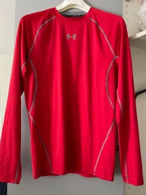 Röd långärmad träningströja Under Armour, XL - Snygg röd långärmad träningströja från Under Armour med gråa kontrastsömmar och logga på bröstet. Tillverkad i ett lätt och stretchigt funktionsmaterial som andas, perfekt för löpning eller gym. Tight passform och sportig design.