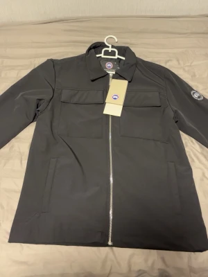 Svart rock från Canada Goose - Stilren svart rock från Canada Goose med rak passform, klassisk krage och två bröstfickor med lock. Rocken har hel dragkedja framtill och en diskret logotyp på ärmen. Materialet är slätt och tåligt, perfekt för kyligare dagar.