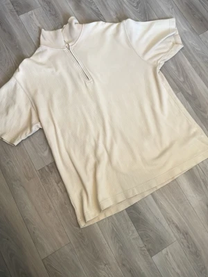 Beige polotopp med half zip - Snygg beige polotopp med kort ärm och half zip framtill. Toppen har en diskret ribbad struktur och hög krage som ger en clean look. Perfekt att styla med jeans eller kjol för en trendig vibe.