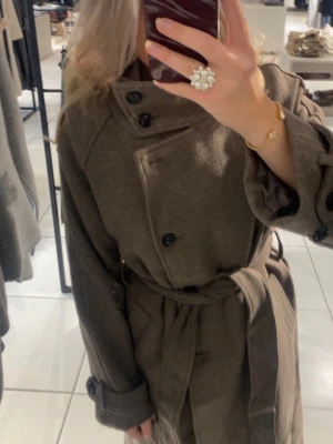 Brun trenchcoat med knytskärp - Stilren brun trenchcoat med dubbelknäppning och knytskärp i midjan. Jackan har stora svarta knappar, bred krage och långa ärmar med knappdetaljer vid ärmsluten. Tillverkad i ett tjockare tyg som ger en klassisk look. Från Gina 