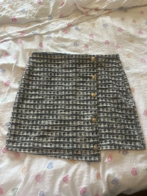 Rutigt kjol från Vero Moda - Kort kjol från Vero Moda i svartvitt rutigt tyg med dekorativa guldknappar framtill. Kjolen har en rak passform och är gjord i ett tjockare, strukturerat material som ger en trendig look. Perfekt att matcha med en enkel topp eller stickad tröja.