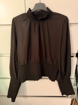 Svart blus med smock från Gina Tricot - Snygg svart blus från Gina Tricot med smockdetaljer vid hals, midja och ärmslut. Den är knappt aldrig använd. Står att det är storlek L men den passar absolut mig som har S vanligtvis.