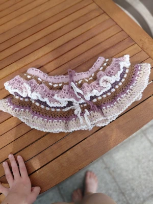 Handvirkad kjol - Supersöt handvirkad kjol i mjuka pastellfärger. Kjolen har volangkant och knytband framtill. Färgkombinationen består av lila, beige, vitt och brunt. Perfekt accessoar för att piffa upp en enkel outfit. Materialet är mjukt garn, troligtvis bomull eller akryl.