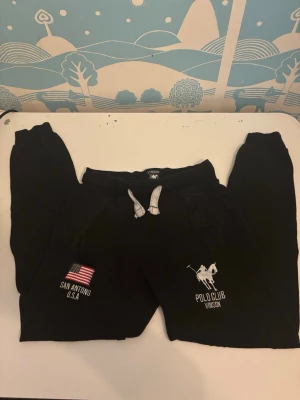 Svarta mjukisbyxor Polo Club Vinson - Svarta mjukisbyxor med vita dragsnören och broderade detaljer. Ena benet har amerikansk flagga och texten 'San Antonio U.S.A', andra benet har Polo Club Vinson-logga med polospelare. Sköna och avslappnade, perfekta för chill dagar.