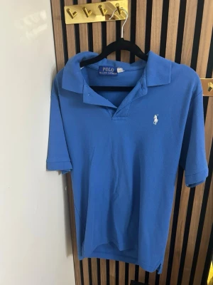 Blå pikétröja från Polo Ralph Lauren - Klassisk blå pikétröja från Polo Ralph Lauren med vit broderad logga på bröstet. Tröjan har korta ärmar, krage och knäppning vid halsen. Perfekt till jeans eller shorts för en avslappnad men snygg look.