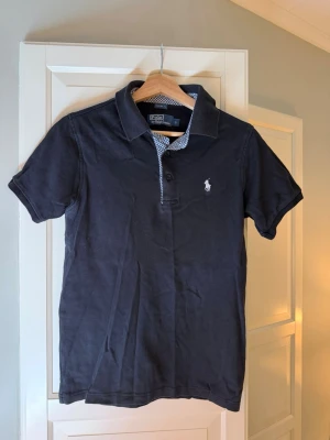 Polo Ralph Lauren piké - Klassisk svart pikétröja från Polo Ralph Lauren med vit broderad logga på bröstet. Tröjan har korta ärmar, krage och knappar framtill. Tillverkad i mjuk bomull och har en stilren look som passar till det mesta.