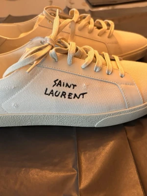 Vita Saint Laurent canvas sneakers/helt nya - Säljer ett par helt nya stilrena sneakers court från Saint Laurent i canvasmaterial med handskriven logga på sidan. Klassisk låg modell med matchande snörning och gummisula. Perfekta för dig som gillar minimalistisk och trendig design.