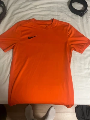 Orange tränings-tshirt från Nike - Snygg orange tränings-tshirt från Nike med svart logga på bröstet. Tillverkad i ett lätt och ventilerande material som passar perfekt till gymmet eller löprundan. Klassisk passform med rund hals och korta ärmar.