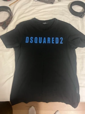 Svart DSQUARED2 t-shirt med blå text - Snygg svart t-shirt från DSQUARED2 med stor blå logga över bröstet. Klassisk passform och rund halsringning. Perfekt till jeans eller shorts för en clean och modern look.