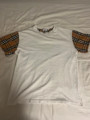 Burberry t-shirt - Säljer en vit t-shirt med klassiska bruna, svarta och vita rutiga kortärmar. T-shirten har en rak passform och är tillverkad i mjuk bomull. Perfekt för dig som vill ha en enkel men ändå unik look.