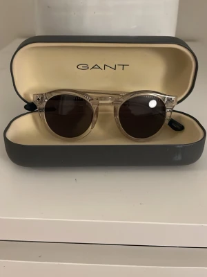 Beige runda solglasögon från Gant - Snygga runda solglasögon från Gant med transparent beige båge och mörka linser. Kommer i originalfodral. Perfekta för soliga dagar och ger en cool vibe till din outfit.