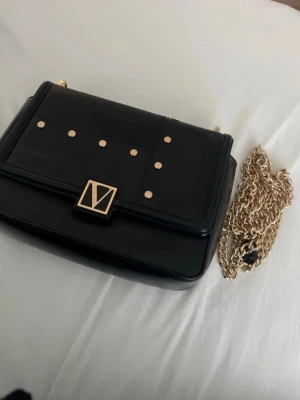 Svart axelväska från Victoria's Secret - Snygg svart axelväska från Victoria's Secret med guldfärgade detaljer och kedjerem. Väskan har dekorativa nitar på framsidan och ett elegant V-emblem i guld. Perfekt storlek för det viktigaste och ger en lyxig touch till din outfit.