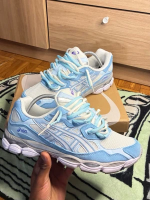  Nya oandvända ASICS sneakers i blått och vitt - Snygga ASICS sneakers i ljusblått och vitt med mesh och mockadetaljer. Skorna har en sportig siluett, tjock sula och klassiska ASICS-loggor på sidan. Perfekta för dig som gillar streetwear och vill ha en fräsch look.