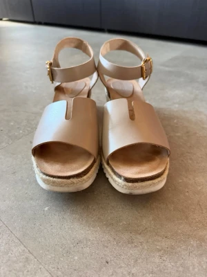 Beige platåsandaler med kilklack - Snygga beige sandaler från Sweeks med breda remmar i skinnimitation och justerbart ankelband med guldfärgat spänne. Sandalerna har platåsula i rep och vit räfflad yttersula samt öppen tå och kilklack för extra höjd.