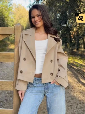 Kort beige trenchcoat med stora knappar - Snygg kort trenchcoat i beige med dubbelknäppning och stora bruna knappar. Jackan har bred krage, långa ärmar och justerbara remmar vid ärmsluten. Perfekt för dig som vill ha en trendig och avslappnad look.