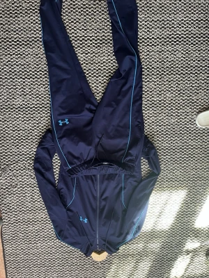 Mörkblå träningsdress Under Armour - Snygg mörkblå träningsdress från Under Armour med ljusblå detaljer och logga på både jacka och byxor. Setet har dragkedja framtill på jackan och smala passformer med kontrastfärgade sömmar. Perfekt för träning eller chill.