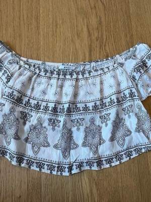 Vit offshoulder topp Gina Tricot - Säljer en vit offshoulder topp från Gina Tricot med svart, detaljerat mönster. Toppen har korta ärmar och resår upptill för en snygg passform. Perfekt för sommarens alla tillfällen och ger en bohemisk vibe.
