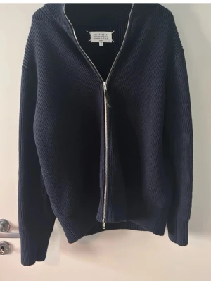 Maison margiela tröja  - Mörkblå stickad tröja med dragkedja framtill och ribbade muddar. Tröjan har en avslappnad passform och lång ärm, perfekt för lager på lager. Enkel och stilren design som passar till det mesta.