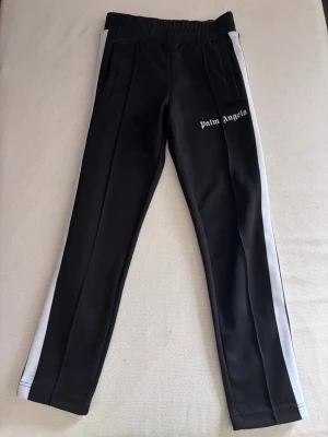 Palm Angels byxor - Palm Angels svarta mjukisbyxor med vita sidoränder och logga på vänster ben. Byxorna har resår i midjan, raka ben och dragkedjor vid bensluten. Tillverkade i ett mjukt och stretchigt material som ger en sportig look. Byxorna är i ett väldigt bra skick!🙂 nypris: 2500kr