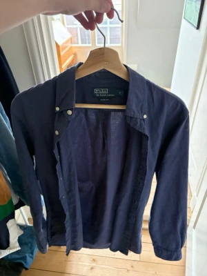 Mörkblå skjorta från Polo Ralph Lauren - Klassisk mörkblå skjorta från Polo Ralph Lauren med knappar hela vägen och lång ärm. Skjortan har en tidlös design med button-down krage och är tillverkad i mjuk bomull. Perfekt för en clean och stilren look.