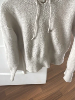 Beige fluffig hoodie med huva - Mysig beige hoodie med fluffig textur och dragsko i huvan. Hoodien har långa ärmar och ribbade muddar vid ärmslut och nederkant. Perfekt för dig som gillar en avslappnad och bekväm stil.