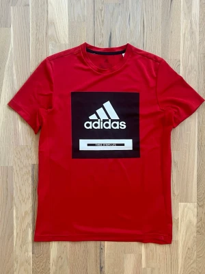 Röd Adidas t-shirt med logga - Snygg röd t-shirt från Adidas med stor svartvit logga på bröstet. Tillverkad i ett lätt och funktionellt material som passar perfekt till träning. Klassisk passform och korta ärmar.