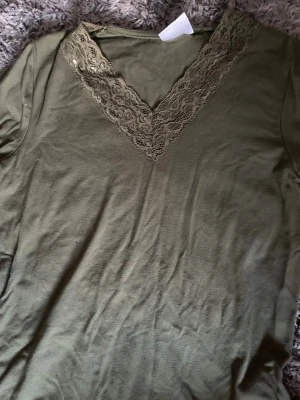 Olivgrön topp med spetsdetalj - Snygg olivgrön topp med v-ringning och dekorativ spets längs halsringningen. Tillverkad i mjukt material och har korta ärmar. Från Gina tricot i storlek XXS