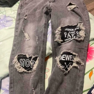 Grå jeans med slitningar och tryck - Säljer ett par grå jeans med coola slitningar och tryckdetaljer. De har en modern, distressed look med texttryck på lår och knän. Perfekta för en avslappnad och trendig stil.  Priset kan sänkas vid snabbt affär