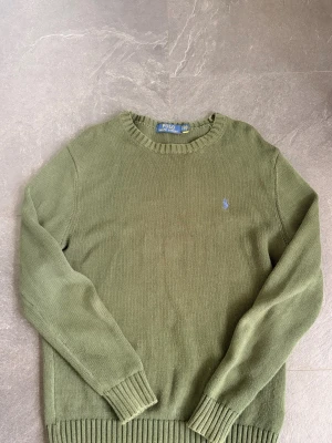 Olivgrön stickad tröja Polo Ralph Lauren - Snygg olivgrön stickad tröja från Polo Ralph Lauren med rund halsringning och ribbade muddar. Tröjan har en liten broderad logga i blått på bröstet och är tillverkad i ett mjukt stickat material. Perfekt för lager på lager och enkel att matcha.