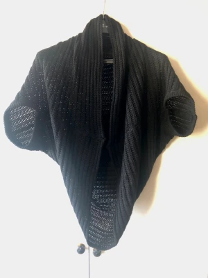 Svart Stickad Cocoon-kofta / Cape – Onesize - Snygg svart stickad kofta i draperad cocoon-/capemodell med fint hålmönster. Går ner över ryggen, har korta fladdermusärmar och är helt öppen framtill för ett väldigt fint fall. Saknar märke, material- och storlekslapp, men uppskattas som en skön akryl/bomullsblandning. Flexibel i storleken och passar Onesize (ca S–L beroende på önskad passform). Fint begagnat skick!