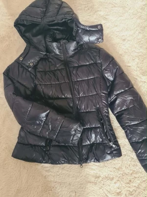Svart pufferjacka  - Snygg svart pufferjacka i glansigt material. Jackan har avtagbar huva, dragkedja och två fickor med dragkedja. Perfekt för kalla höstdagar💕 strl xxs 