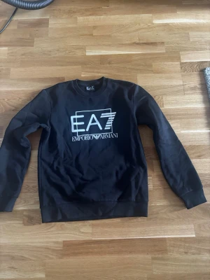 Svart sweatshirt från Emporio Armani - Snygg svart sweatshirt från Emporio Armani EA7 med stor vit logga framtill. Tröjan har rund halsringning, långa ärmar och ribbade muddar vid ärmslut och nederkant. Perfekt för en stilren och avslappnad look. Helt ny används 1 gång.