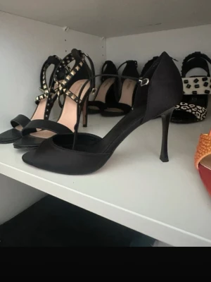 Svarta pumps med öppen tå från Mango - Stilrena svarta pumps från Mango med öppen tå och smal klack. Skorna har en elegant rem runt ankeln och är tillverkade i ett matt tygmaterial. Perfekta för att lyfta vilken outfit som helst. Oanvända i storlek 38