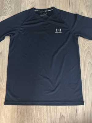Långärmad Under armour träningströja - Svart långärmad träningströja från Under Armour i funktionsmaterial. Tröjan har en diskret logga på bröstet och Under Armour-tryck längs ryggen. Perfekt för gym, löpning eller annan träning. OBS: färgen är väldigt mörkblå.