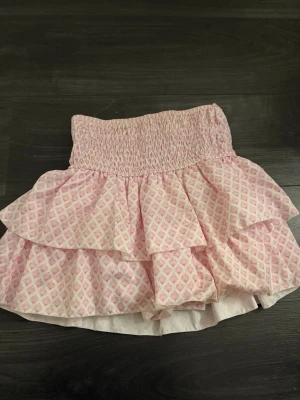 Rosa volangkjol från Meet x Tiger - Supersöt kort volangkjol från Meet x Tiger i rosa och vitt med smock i midjan. Med inbyggda shorts. Kjolen har ett diskret rutmönster och dubbla volanger som ger en lekfull look. Perfekt för vår och sommar!