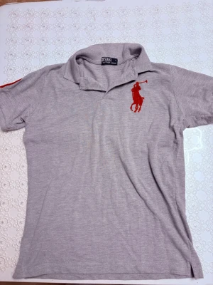 Grå piketröja/Polo Ralph Lauren - Säljer en klassisk grå pikétröja från Polo Ralph Lauren med röd broderad logga på bröstet. Tröjan har korta ärmar, krage och är gjord i mjuk bomull. Perfekt till en avslappnad men ändå stilren look.