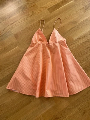  Ny trendig  orange kortklänning från Zara  - Söt kortklänning i orange med tunna vita ränder och smala axelband. Klänningen har en lätt A-linjeformad siluett och är perfekt för varma dagar. Tillverkad i ett mjukt bomullstyg som känns skönt mot huden.