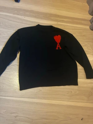 Svart stickad tröja med rött hjärta - Svart stickad tröja med ett rött hjärta och bokstaven A på bröstet. Tröjan har rund halsringning och långa ärmar. Perfekt för dig som gillar stilrena plagg med en twist. Materialet ser ut att vara mjukt och bekvämt.