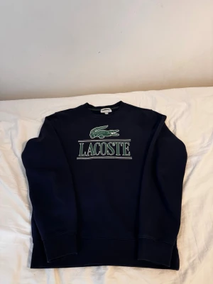 Mörkblå sweatshirt från Lacoste - Snygg mörkblå sweatshirt från Lacoste med stort grönt och vitt logotryck på bröstet. Klassisk rund halsringning och långa ärmar. Perfekt för dig som gillar streetwear och vill ha en stilren tröja med tydlig logga.