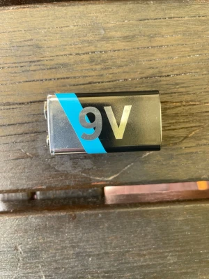 9V batteri med blå detalj - Säljer ett klassiskt 9V batteri med svart och silver färg samt en blå diagonal detalj. Perfekt till elektronikprylar som behöver extra kräm. Batteriet har standardform och metallhölje.