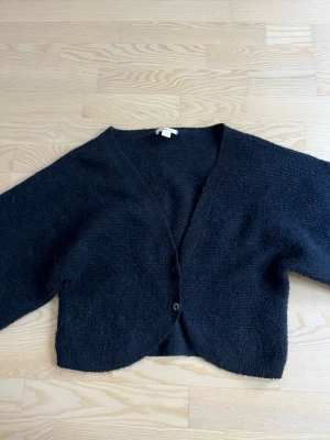 Svart fluffig kofta med v-ringning - Mysig svart kofta med v-ringning och två knappar framtill. Koftan har en fluffig textur och är något croppad i modellen. Perfekt att slänga över en t-shirt eller linne för en chill look.