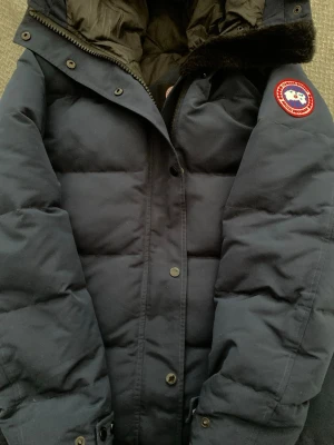 Mörkblå dunjacka från Canada Goose - Säljer en mörkblå dunjacka från Canada Goose med klassisk patch på ärmen. Jackan har hög krage, dragkedja och knappar framtill samt en varm, vadderad design och detaljer i svart fuskpäls vid huvan. Perfekt för kalla vinterdagar.