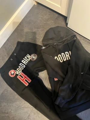 Svart hoodie med HOODRICH-tryck - Hoodrich sett! Tröjan är m och byxor S. Svart super sköna kläder .hoodie med stor vit HOODRICH-text på bröstet, röda och vita patchar samt broderad H-detalj. Hoodien har huva med dragsko och känguruficka framtill. Snygg streetwear-stil med tydliga loggor och coola detaljer.