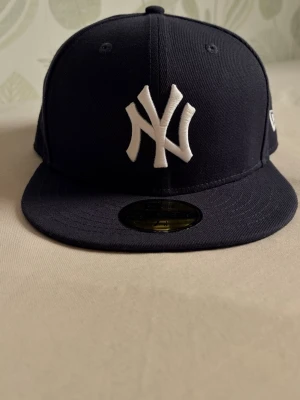 Svart New Era Yankees keps 59FIFTY - Säljer en svart New Era 59FIFTY keps med klassiskt New York Yankees broderat vitt logotyp framtill. Kepsen har rak skärm och sticker ut med sin streetwear-vibe. Tillverkad i slitstarkt tyg och har ventilationhål upptill.