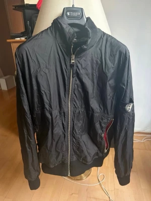 Svart vindjacka från Stone Island storlek L äkta  - Snygg svart vindjacka från Stone Island med dragkedja framtill, hög krage och ribbade muddar. Jackan har den klassiska Stone Island-loggan på ärmen och röda detaljer vid fickan. Tillverkad i lätt, glansigt material som passar perfekt till streetwear. Köpt 2015 så den är gammal med knappt använd . Äkta 