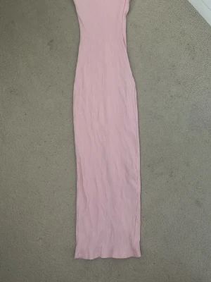 Lång rosa ribbad klänning - Lång, tight klänning i ljusrosa ribbat material. Klänningen är ärmlös och har en slits baktill för extra rörelsefrihet. Perfekt för dig som gillar en enkel men stilren look.