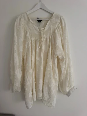 Vit/creme långärmad klänning från Boohoo - Luftig vit klänning från Boohoo med boho-vibbar. Klänningen har långa, lösa ärmar, knytning vid halsen och ett diskret blommigt mönster i tyget. Perfekt för sommarens alla tillfällen och ger en lyxig känsla.