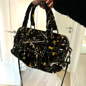 Zadig & Voltaire väska - Cool svart handväska från Zadig & Voltaire med färgstänk i vitt, gult och vinrött. Väskan har två handtag, avtagbar axelrem och dubbla dragkedjor framtill. Materialet är mocka och den har en silverfärgad logga med vingar på framsidan. Finns inte att köpa på Zadig längre. Pris kan diskuteras 