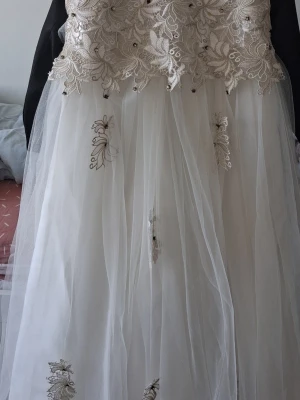 Vit långklänning med spets och rosett - Elegant vit långklänning med broderad spets upptill och transparent tyllkjol. Klänningen har fina blommiga detaljer, en stor rosett i midjan och vackra broderier på kjolen. Perfekt för speciella tillfällen när du vill känna dig extra fin.